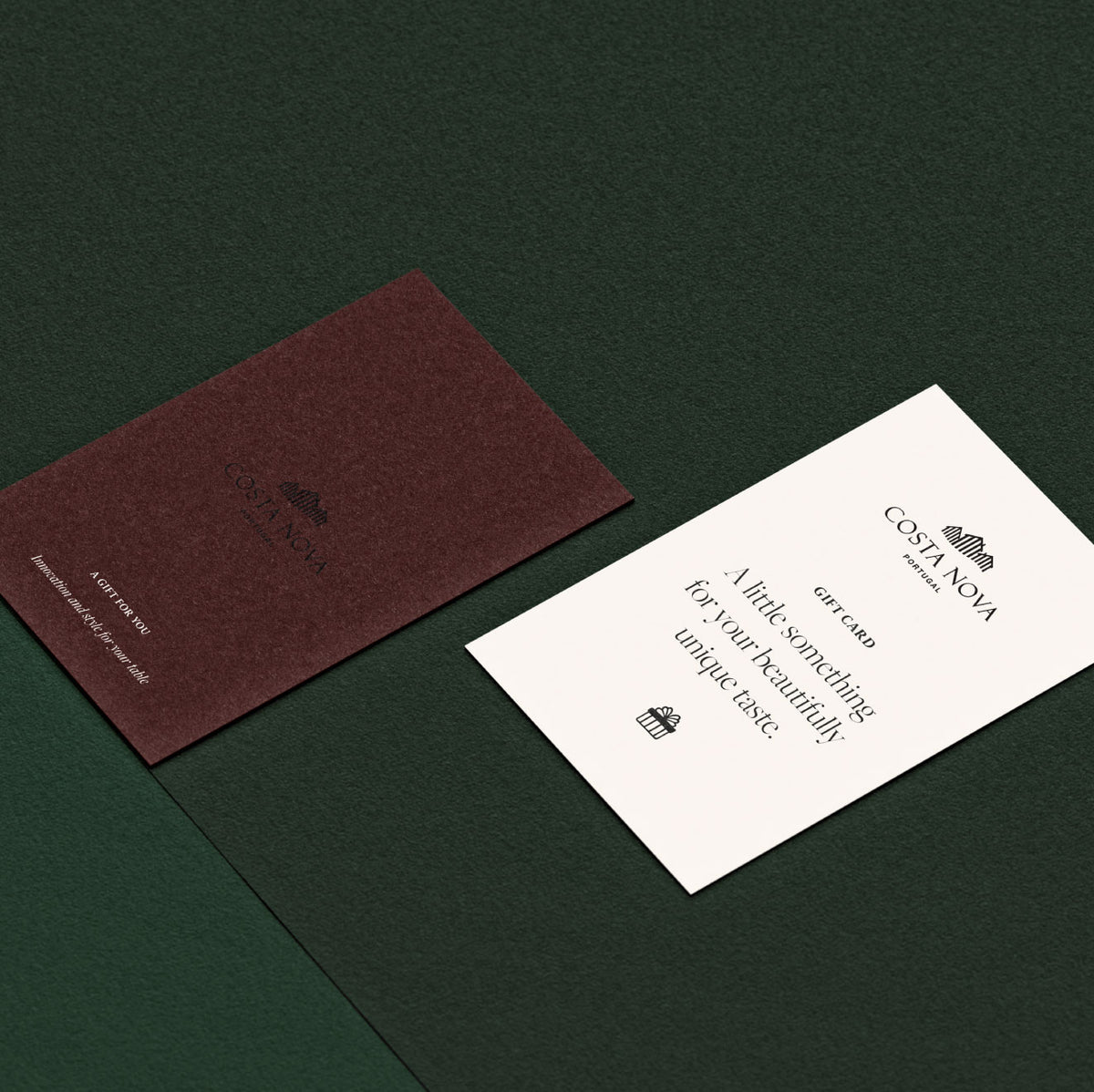 Two Costa Nova gift cards, one maroon and one cream with a small gift icon, rest on a dark green textured surface.